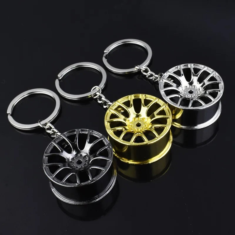 Drei metallische ShineMasters Felgen-Schlüsselanhänger mit Miniatur-Autofelgen-Designs sind in einer Reihe auf einer schwarzen Oberfläche angeordnet. Von links nach rechts sind die Farben der Felgen schwarz, gold und silber. Jeder Schlüsselanhänger hat einen runden Ring und eine Kette, die an der Autofelge befestigt ist – ein ideales Accessoire für jeden Autoliebhaber.
