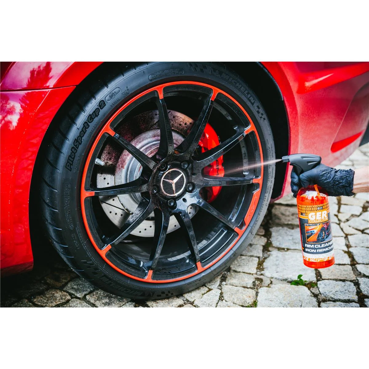 Eine Person mit einem schwarzen Handschuh sprüht mit einem GERcollector RIM CLEANER & IRON REMOVER von GERcollector eine Reinigungslösung auf die Felge eines Sportwagens. Das rote Auto steht auf Kopfsteinpflaster und das Rad ist mit schwarzen und roten Details versehen, die eisenhaltige Verschmutzungen wirksam bekämpfen.