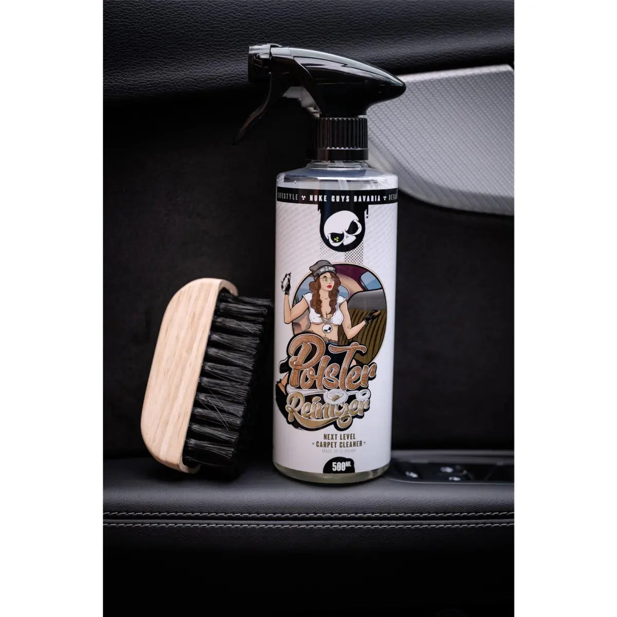 Ein Produktfoto mit dem Nuke Guys Polsterreiniger, einer 500 ml Sprühflasche mit Oberflächenreiniger und mitgeliefertem Sprühkopf, neben einer Bürste mit schwarzen Borsten auf der Innenfläche eines Autos. Die Vintage-Illustration einer Frau auf dem Etikett verstärkt den Reiz und betont, dass es für Textilien und Leder geeignet ist.