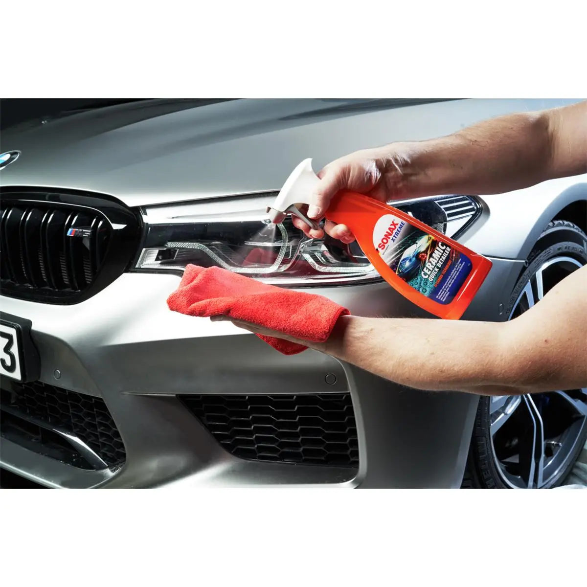 Eine Person reinigt die Scheinwerfer eines silbernen Autos mit einer roten Sprühflasche, die mit Sonax Xtreme Ceramic Quick Detailer 750 ml von SONAX gefüllt ist, und einem roten Mikrofasertuch. Die Scheinwerfer sind klar und die Front des Autos, einschließlich eines Teils des Kühlergrills und der Räder, hat einen spiegelnden Hochglanz.